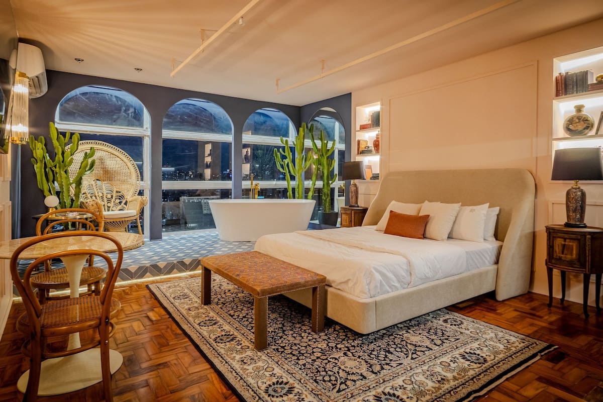 Un dormitorio amplio con una cama grande, una zona de estar con sillones, una mesa de comedor y vistas a través de amplios ventanales.