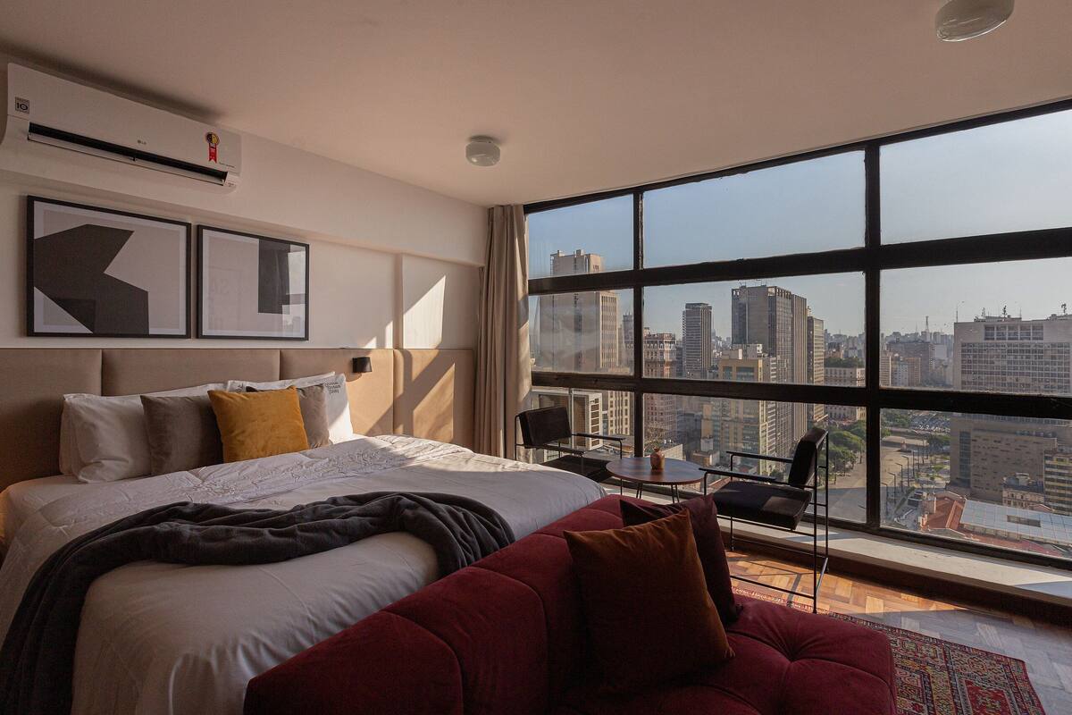 Una habitación de hotel con un ventanal grande, una cama, un sofá rojo y una mesita pequeña.