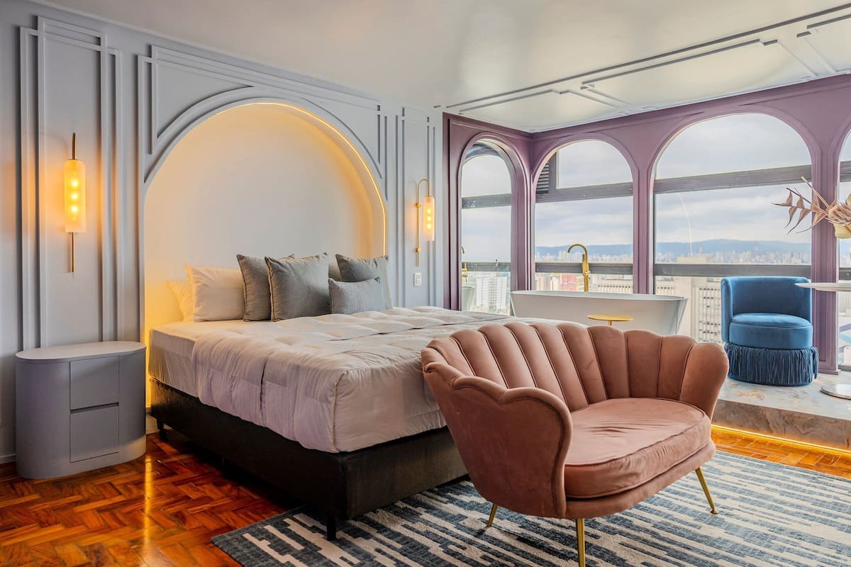 Un dormitorio con cama, una silla, una mesita de noche y vistas a la ciudad a través de amplias ventanas.