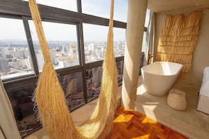 A room with a large window, a bathtub, and a hanging hammock. - Vossa Bossa Sky Mirante (São Paulo)