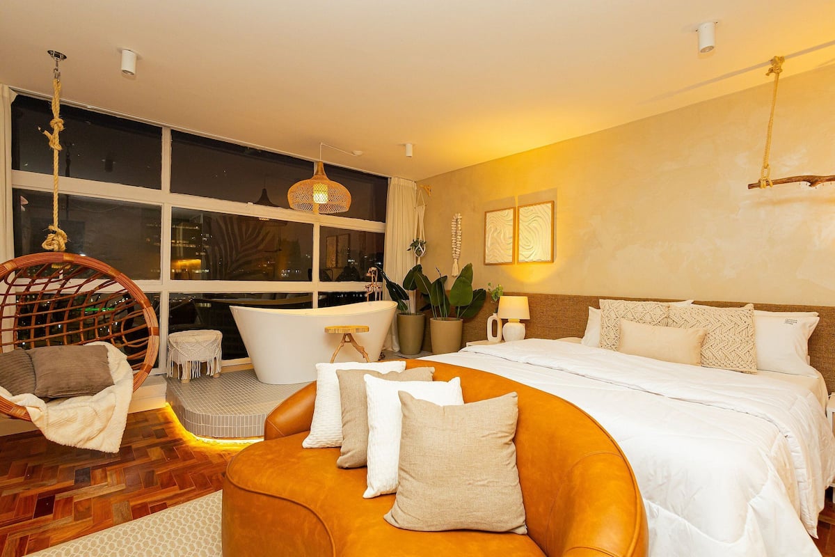 Una habitación de hotel moderna con una cama grande, un sillón colgante, una mesita y vistas de la ciudad de noche.