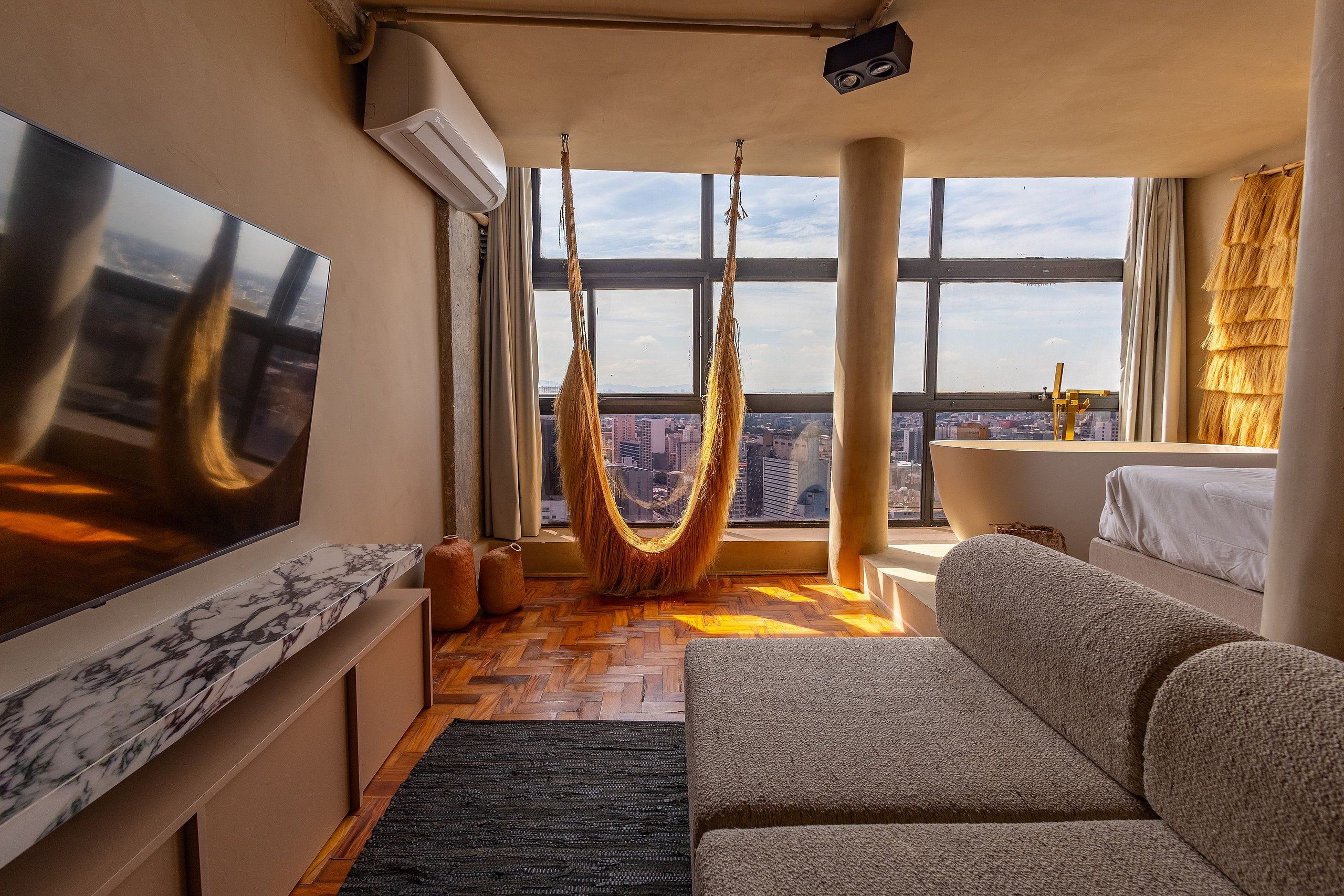 A modern living room with a large TV, a hammock, a sofa, and a view of the city through large windows.