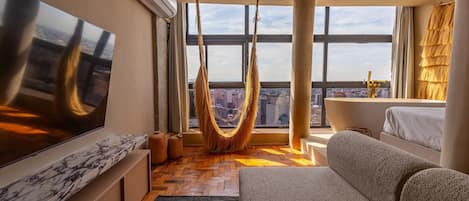 A modern living room with a large TV, a hammock, a sofa, and a view of the city through large windows.
