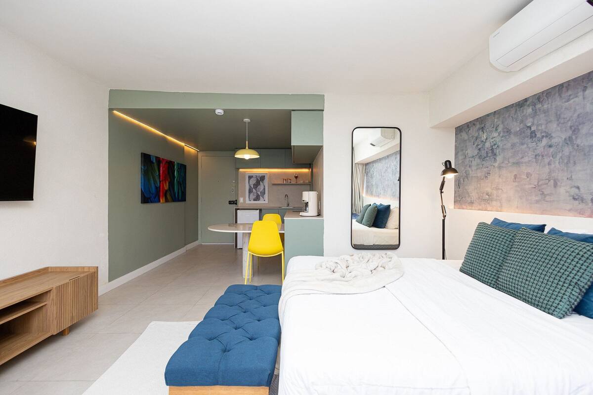 Una habitación de hotel moderna con una cama grande, un escritorio pequeño, una silla amarilla y una kitchenette al fondo.