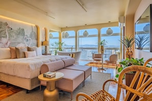 A modern bedroom with a large bed, a sofa, a small table, and a chair. There are plants and a view of the city. - Vossa Bossa Sky Mirante (São Paulo)