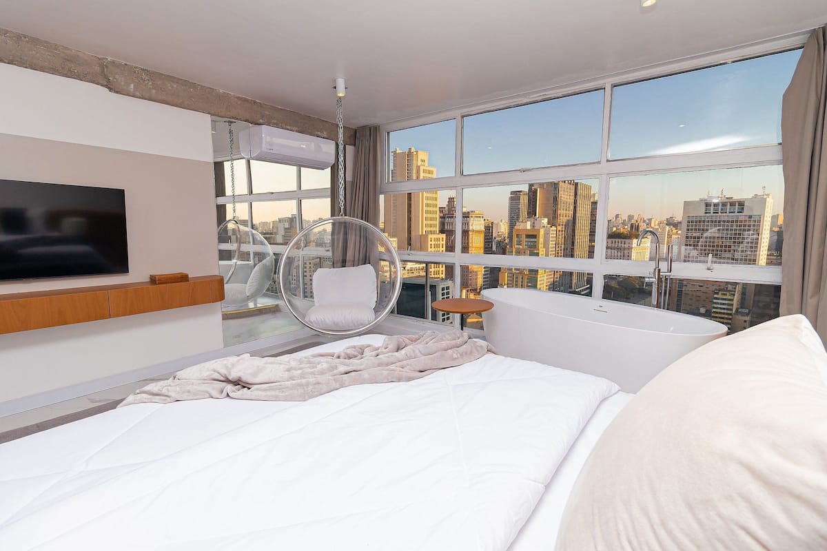 Habitación de hotel moderna con una cama grande, una silla colgante, una bañera y vistas a la ciudad.