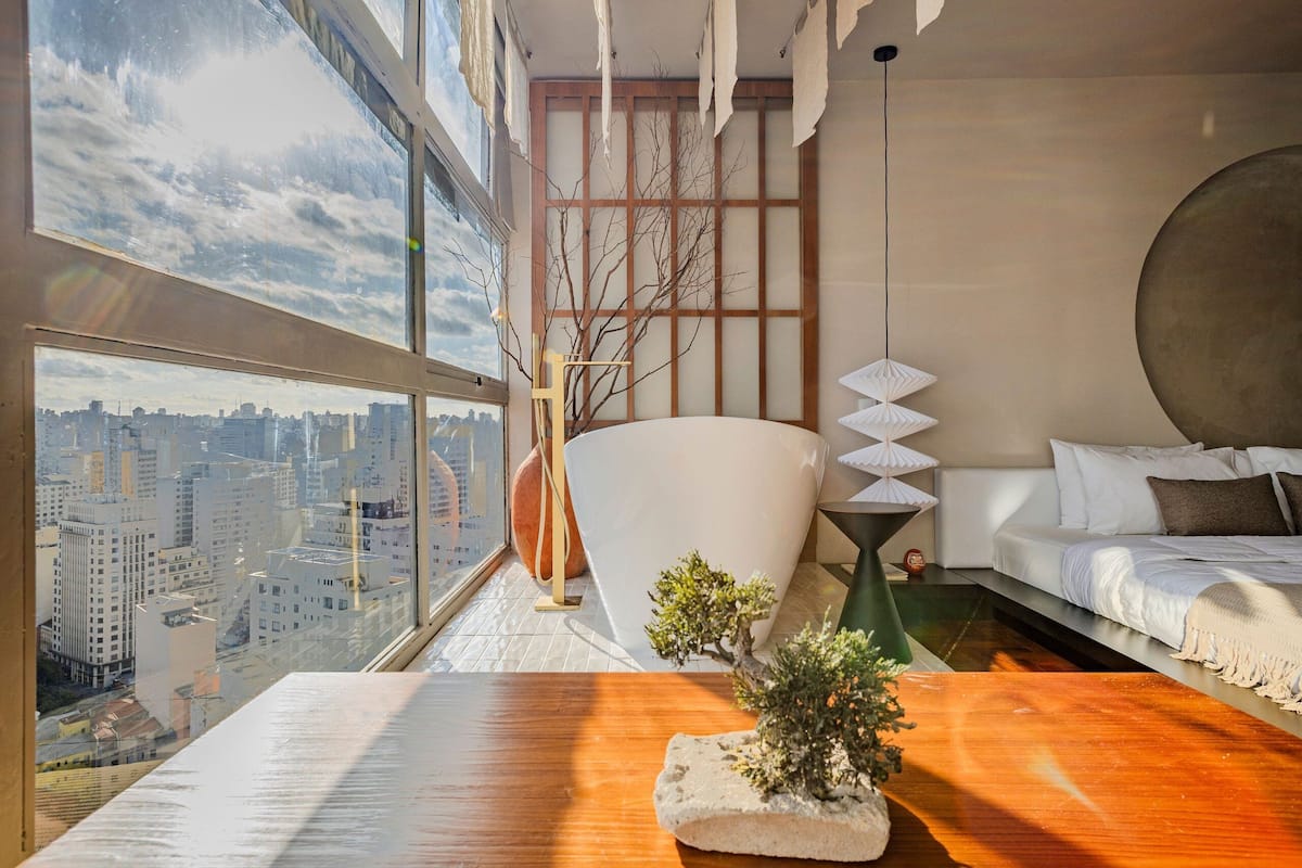 Una habitación de hotel moderna con vista a la ciudad, un ventanal grande, una mesa de madera, un sofá y una planta decorativa.