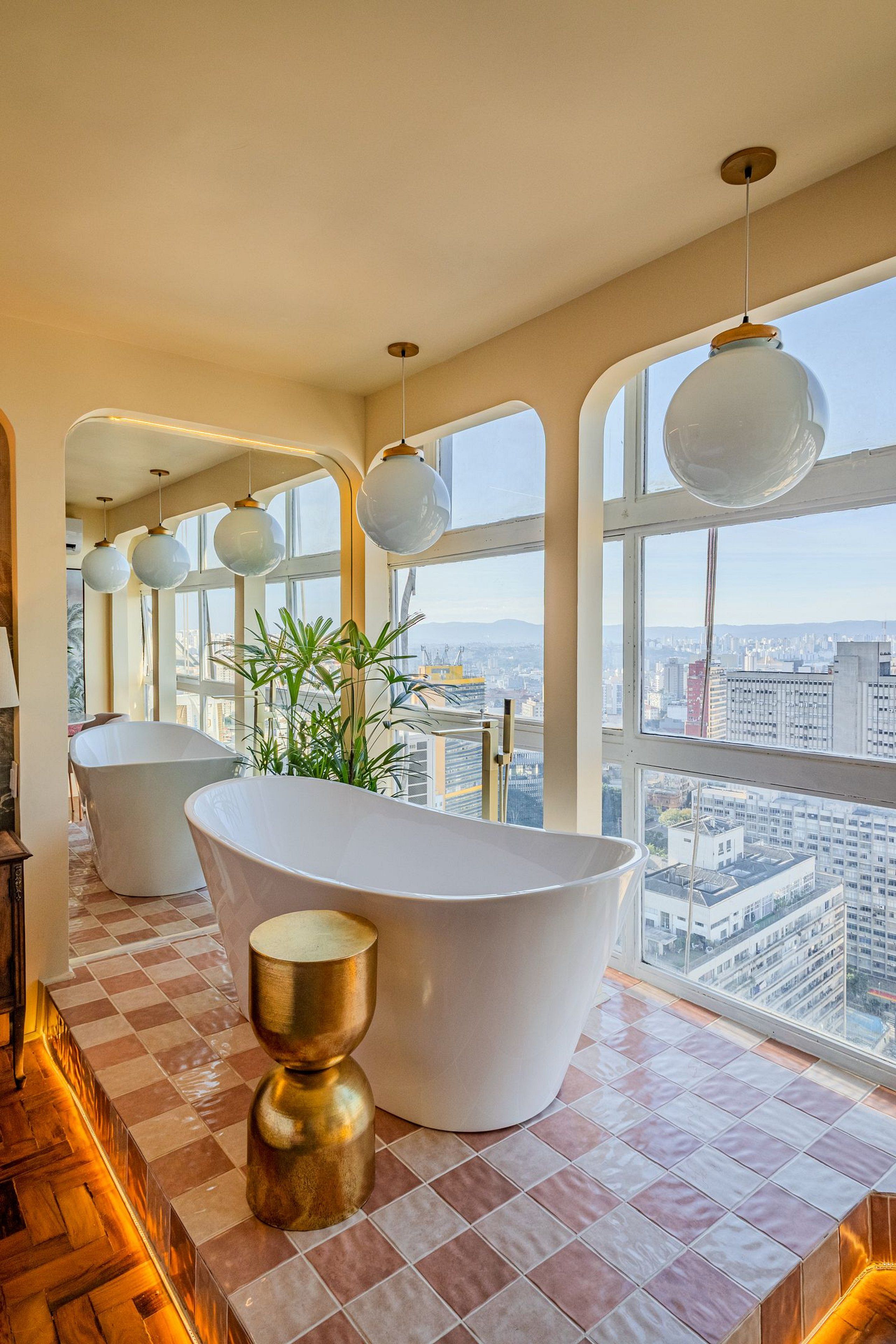 A modern bathroom with a freestanding bathtub, a gold decorative stool, and large windows offering a city view.