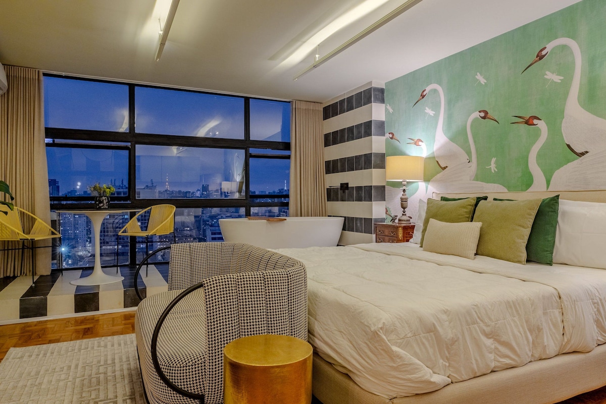 A modern hotel room with a large bed, a view of the cityscape, a dining table, and a chair.