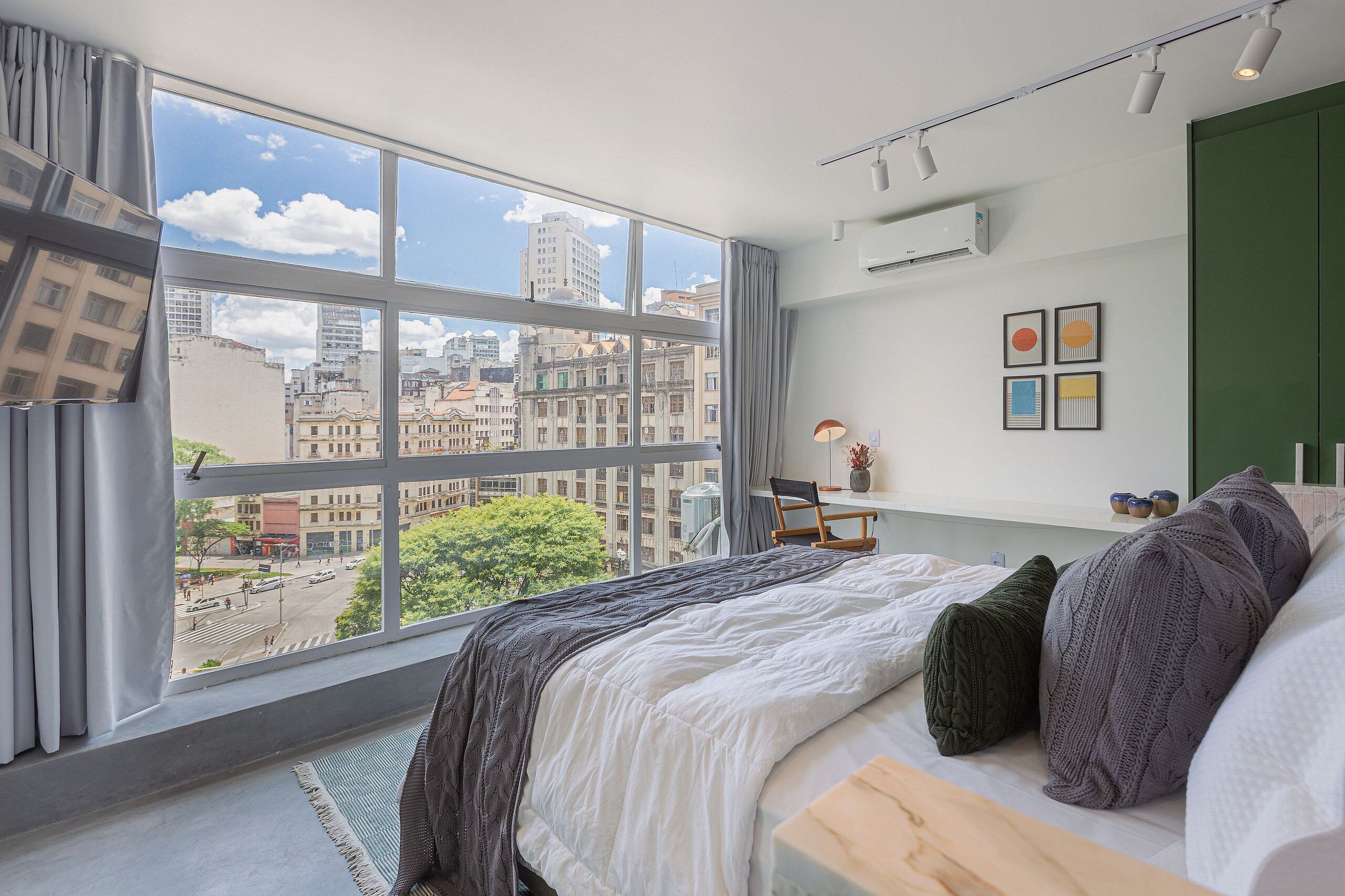 Quarto moderno com uma janela ampla, uma cama, uma escrivaninha e vista para a paisagem urbana.