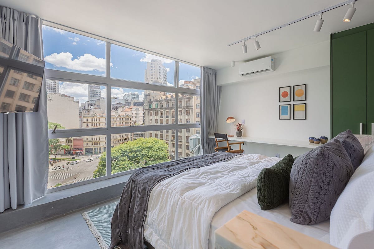 Un dormitorio moderno con un ventanal grande, una cama, un escritorio y vistas a la ciudad.