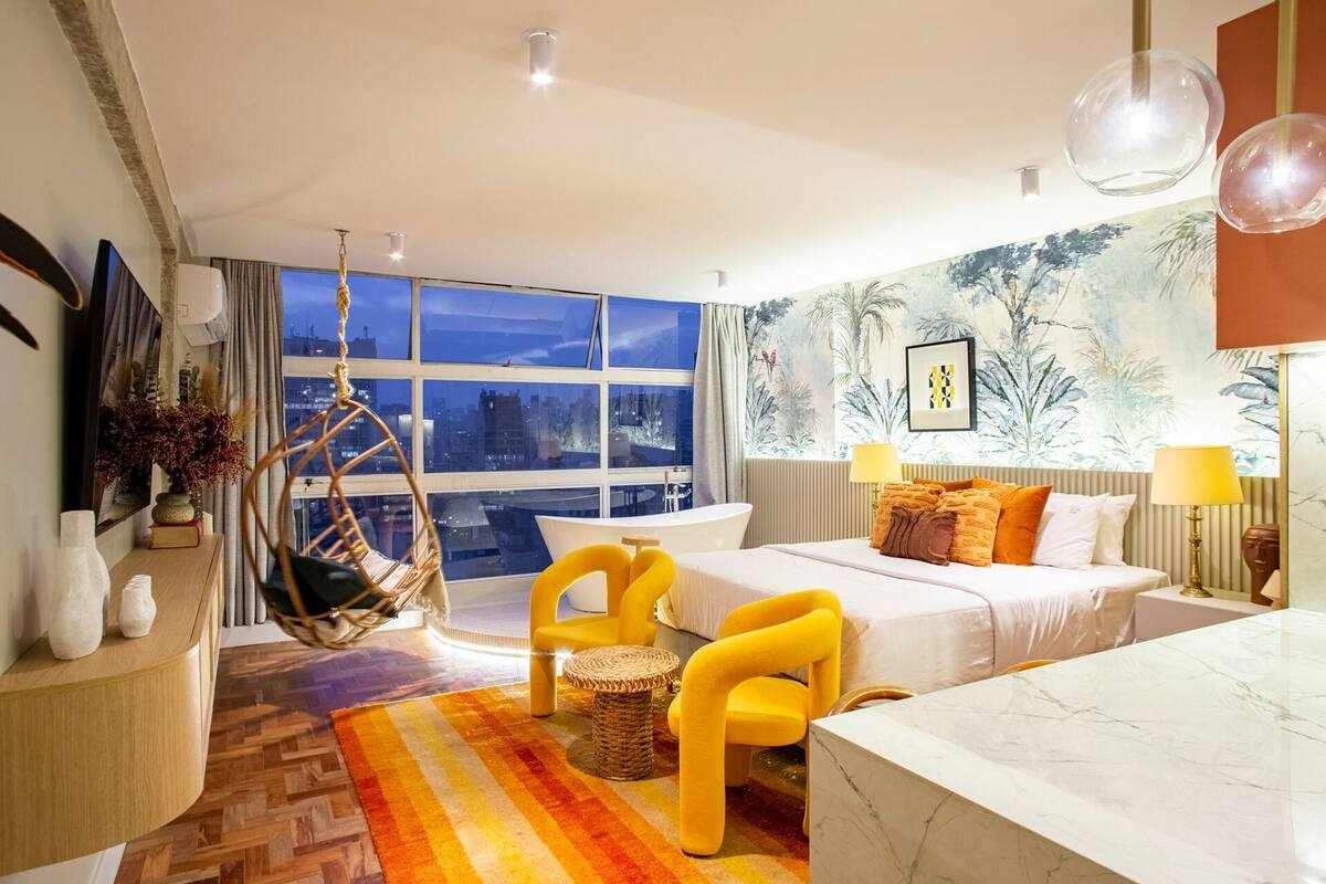 Habitación de hotel moderna con un gran ventanal, una silla colgante, una cama con sillas amarillas, una mesa de mármol y vista a la ciudad.