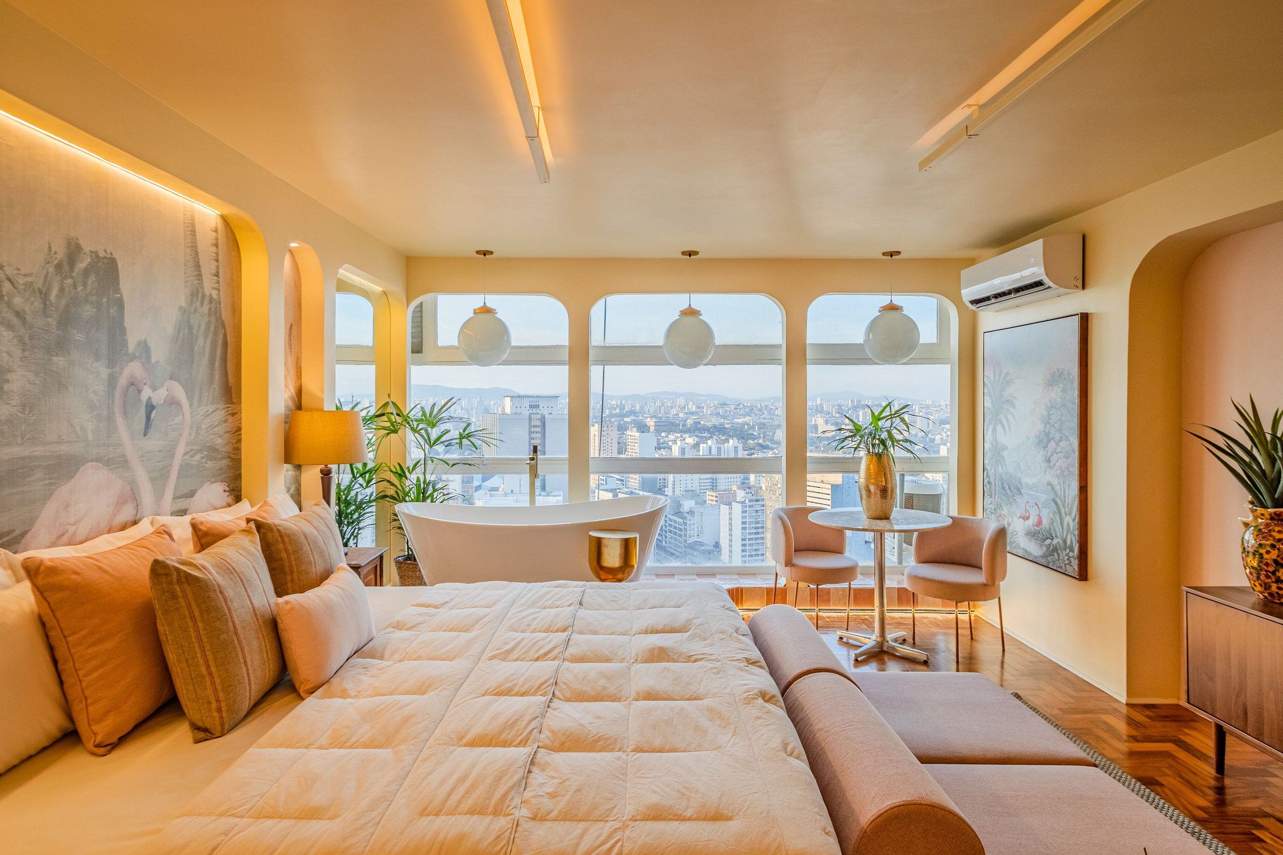 A modern bedroom with a large bed, a sitting area, a bathtub, and a city view.