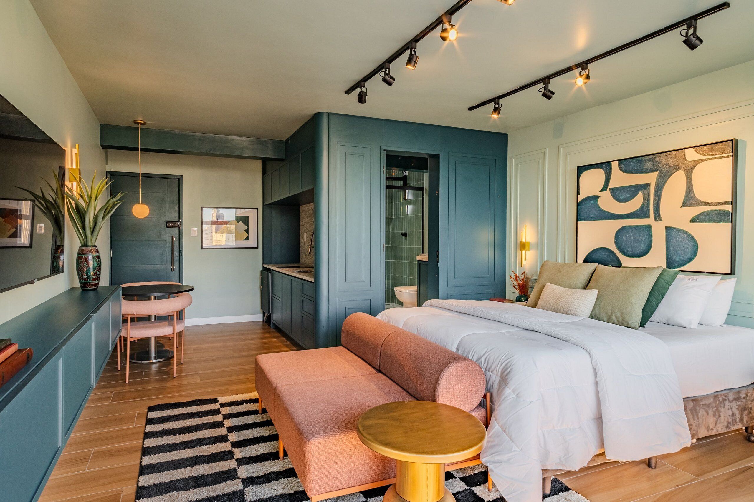 Um quarto moderno com cama, banco, mesa redonda e uma área de cozinha com armários e pia.