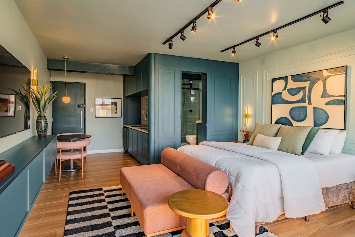 Un dormitorio moderno con cama, banca, mesa redonda y una zona de cocina con armarios y lavabo.