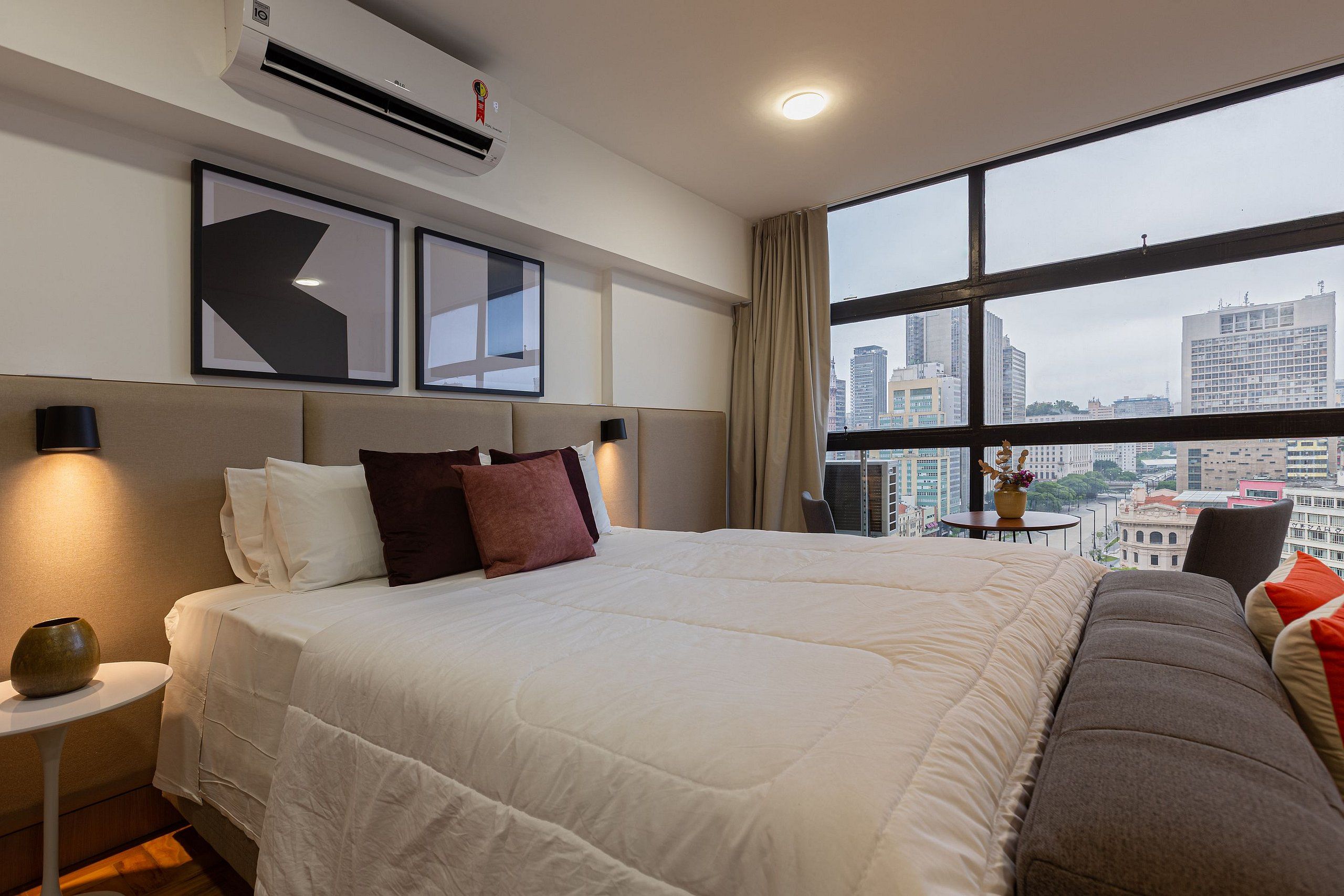 A hotel room with a large bed, a bedside table, a lamp, a view of the city through large windows, and an air conditioning unit.