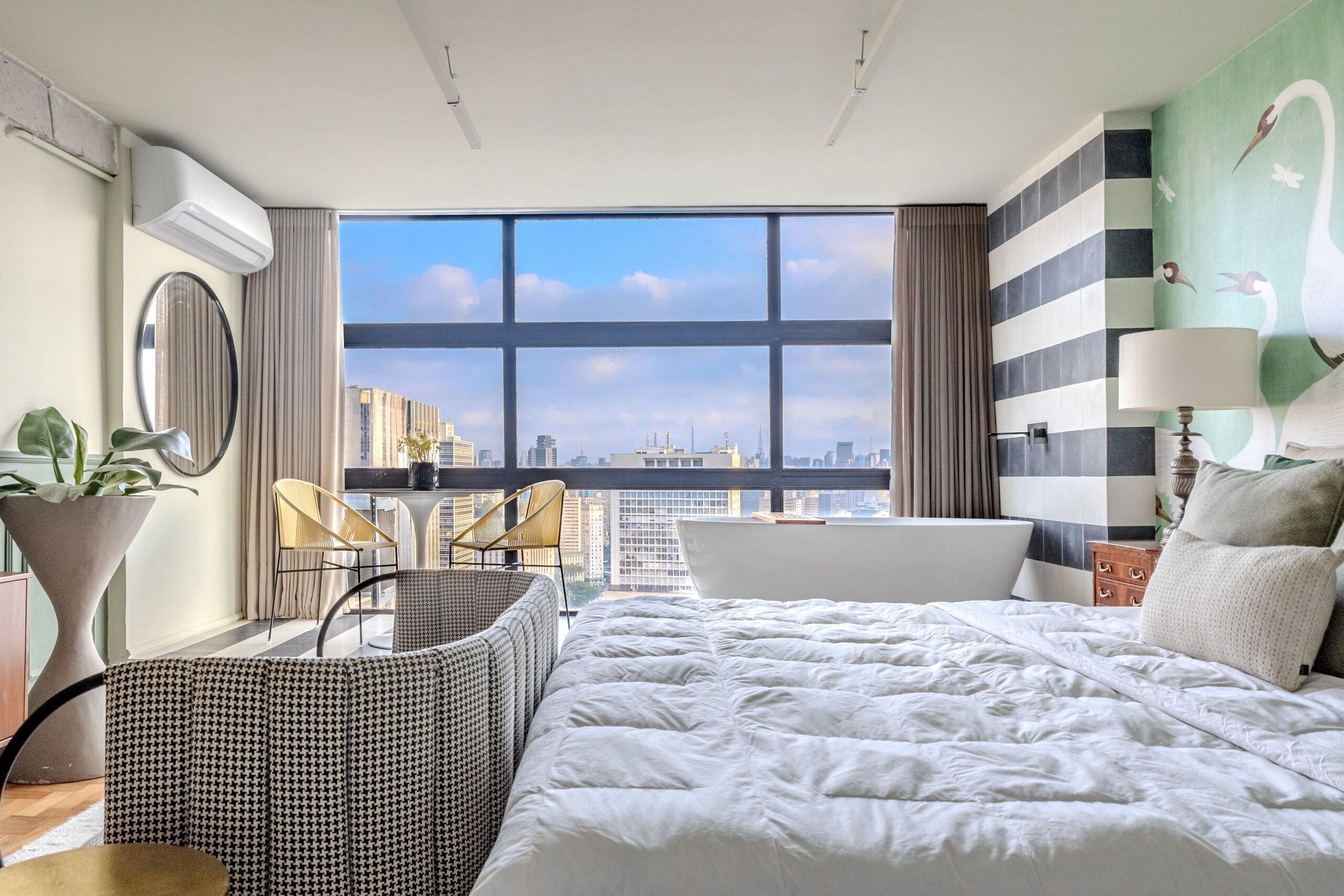 Quarto de hotel moderno com uma cama grande, área de estar com cadeiras e vista para a paisagem urbana, além de uma banheira.