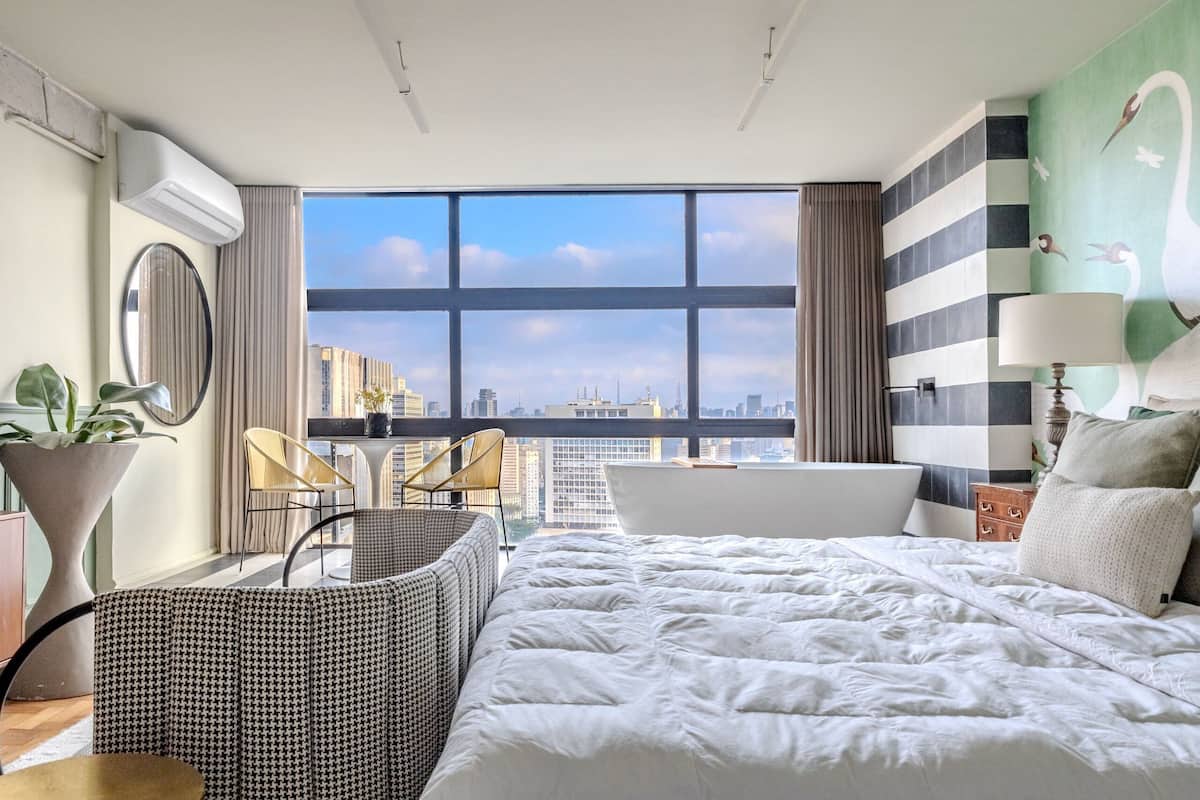 Una habitación de hotel moderna con una cama grande, un área de descanso con sillones, vistas de la ciudad a través de amplios ventanales y una bañera.