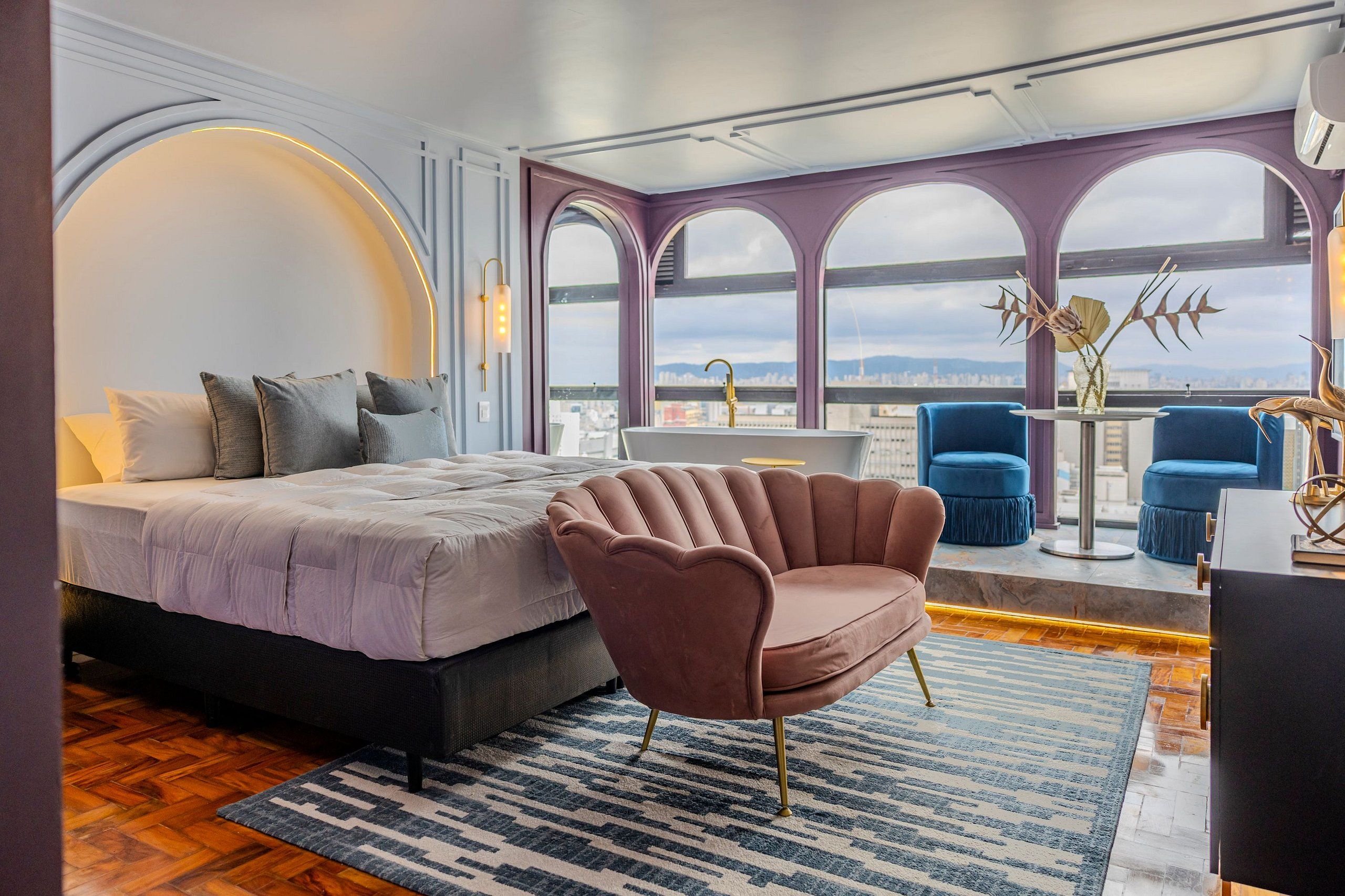 A modern bedroom with a bed, a pink armchair, a blue sofa, a bathroom, and a city view.