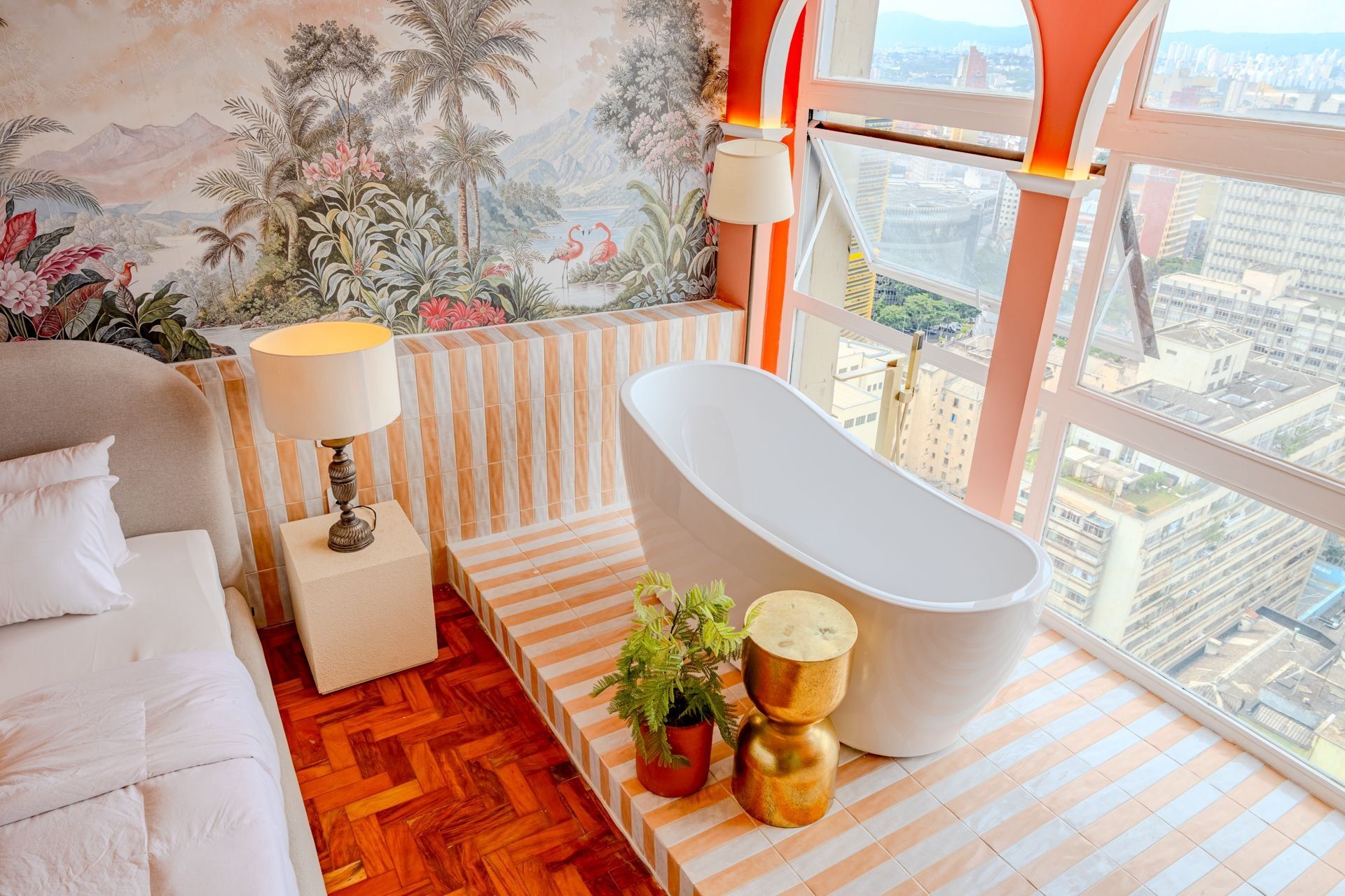 A modern bathroom with a freestanding bathtub, a small bedside table, a lamp, and a potted plant.