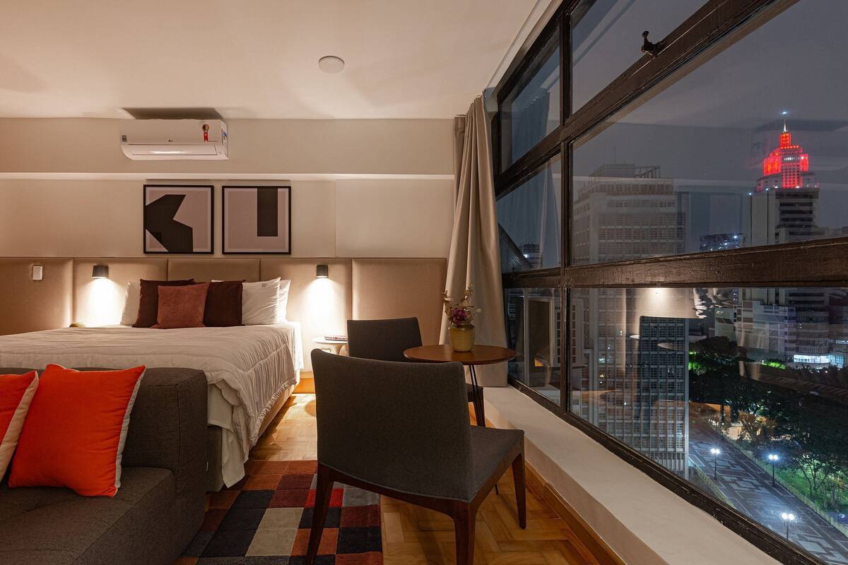 Habitación de hotel con cama, sofá, sillón, una mesita pequeña y un ventanal con vistas a la ciudad.