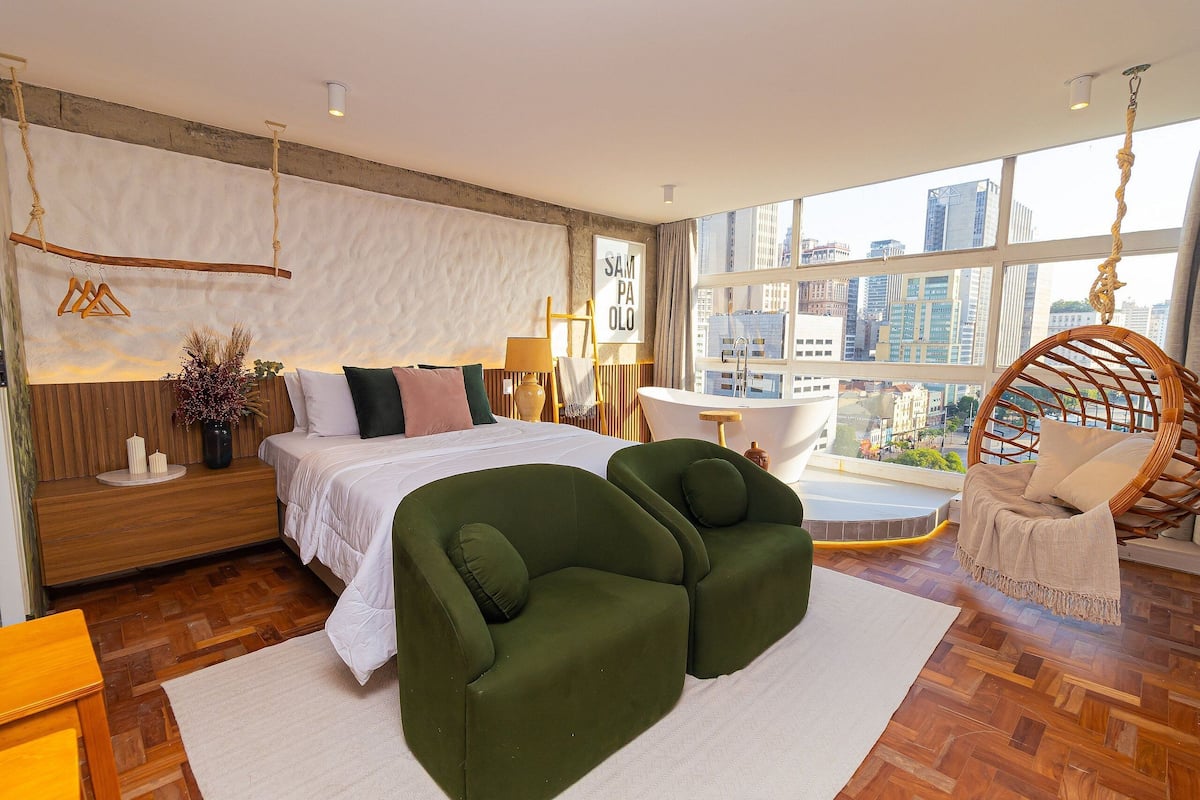 Un dormitorio moderno con una cama grande, dos sillones verdes, un sillón colgante, vistas a la ciudad y un ventanal amplio.