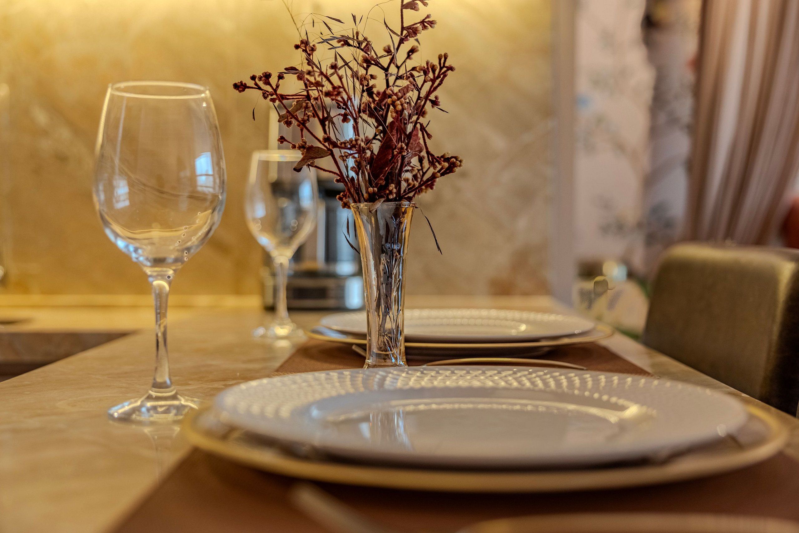 Una mesa de comedor con un jarrón de vidrio transparente que contiene flores secas, una copa de vino y un plato.