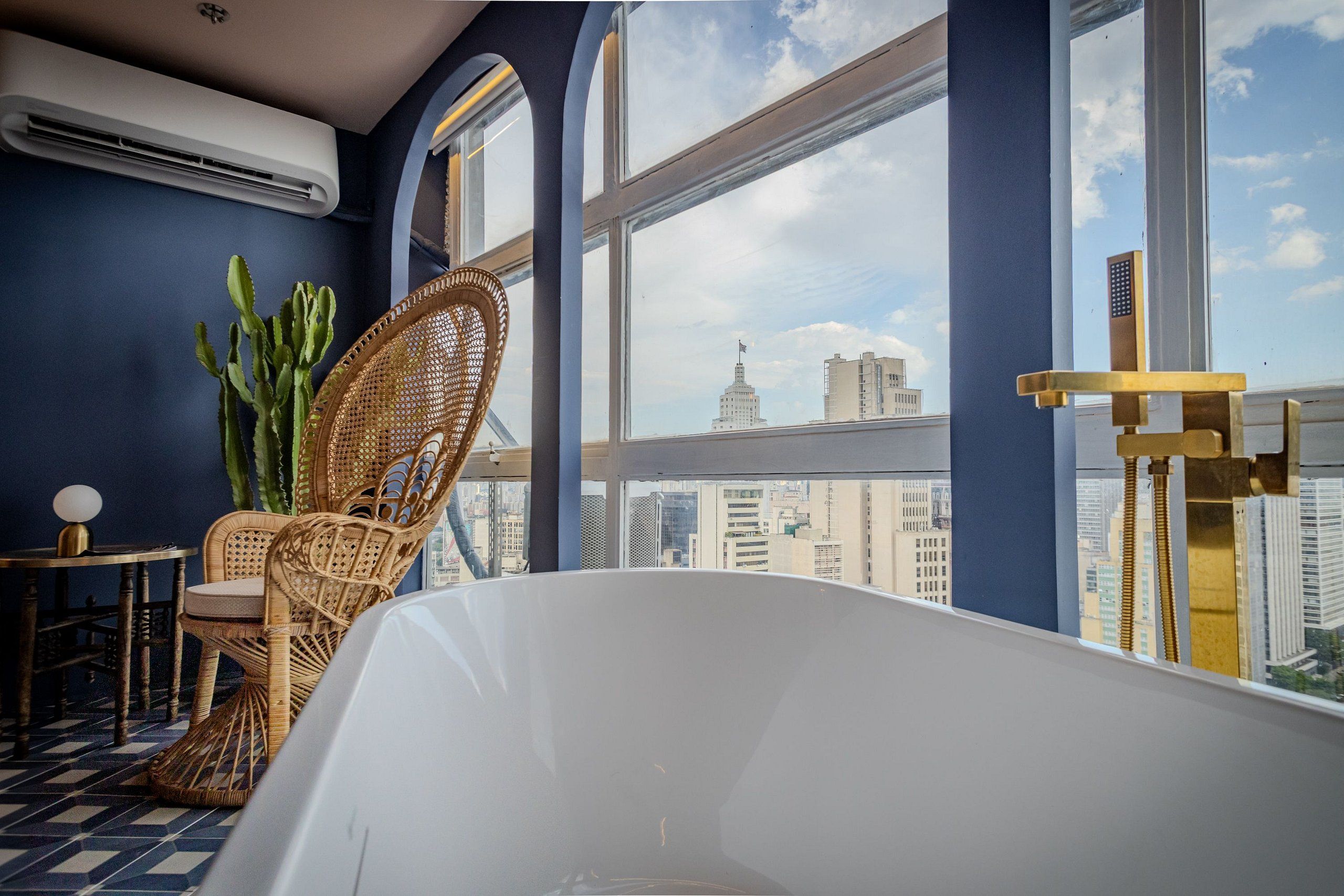 A room with a large bathtub, wicker chairs, a round table, and a view of city buildings through large windows.