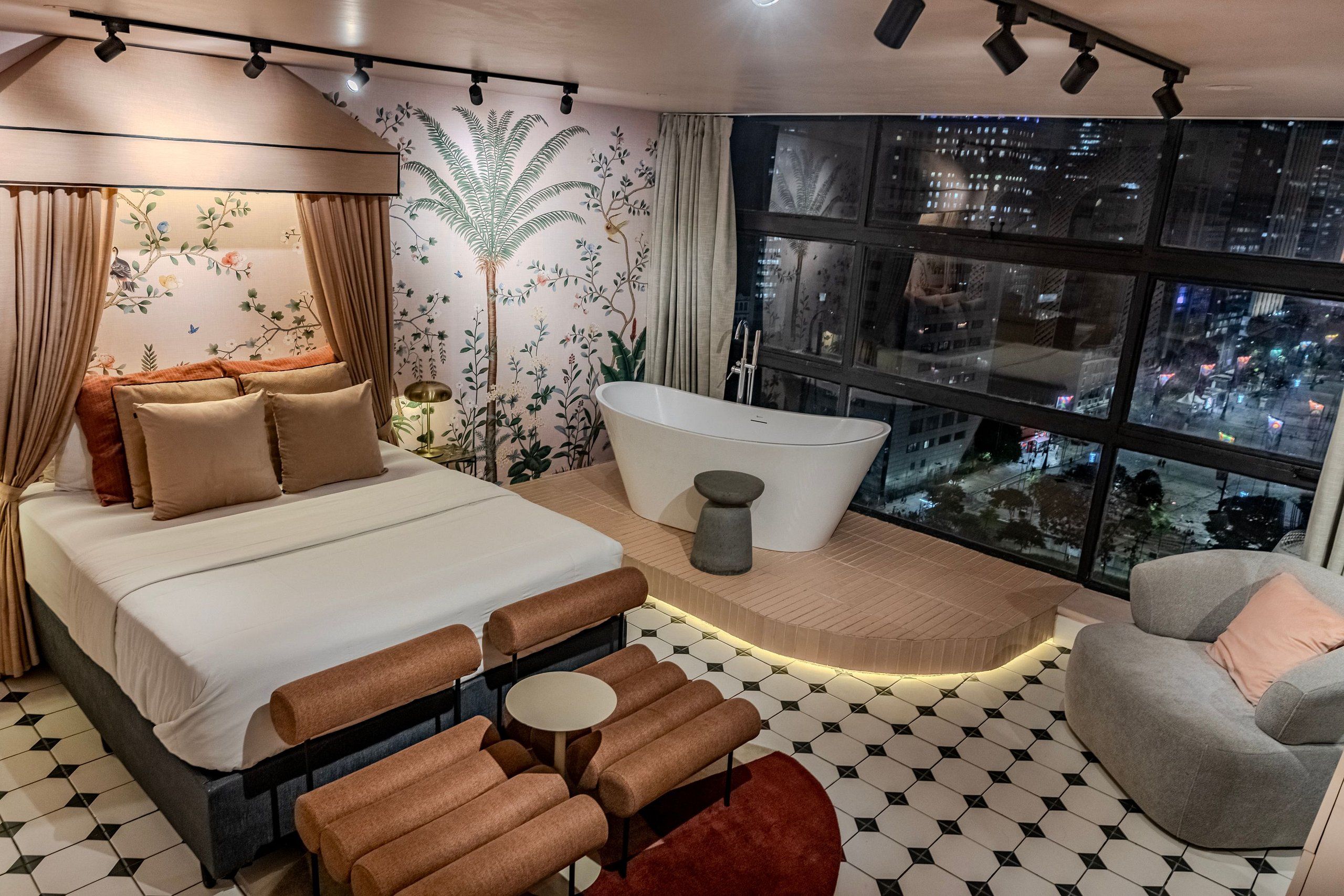Ein modernes Hotelzimmer mit einem großen Bett, einer Sitzecke mit Sofas, einer Badewanne und Blick auf die Stadt durch große Fenster.