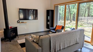 A modern living room with a fireplace, a flat-screen TV mounted on the wall, a sofa, and a chair. - Raccoon Cabin @ Orange Tractor Retreat: a modern cabin on 40 wooded acres (Wisconsin Dells)