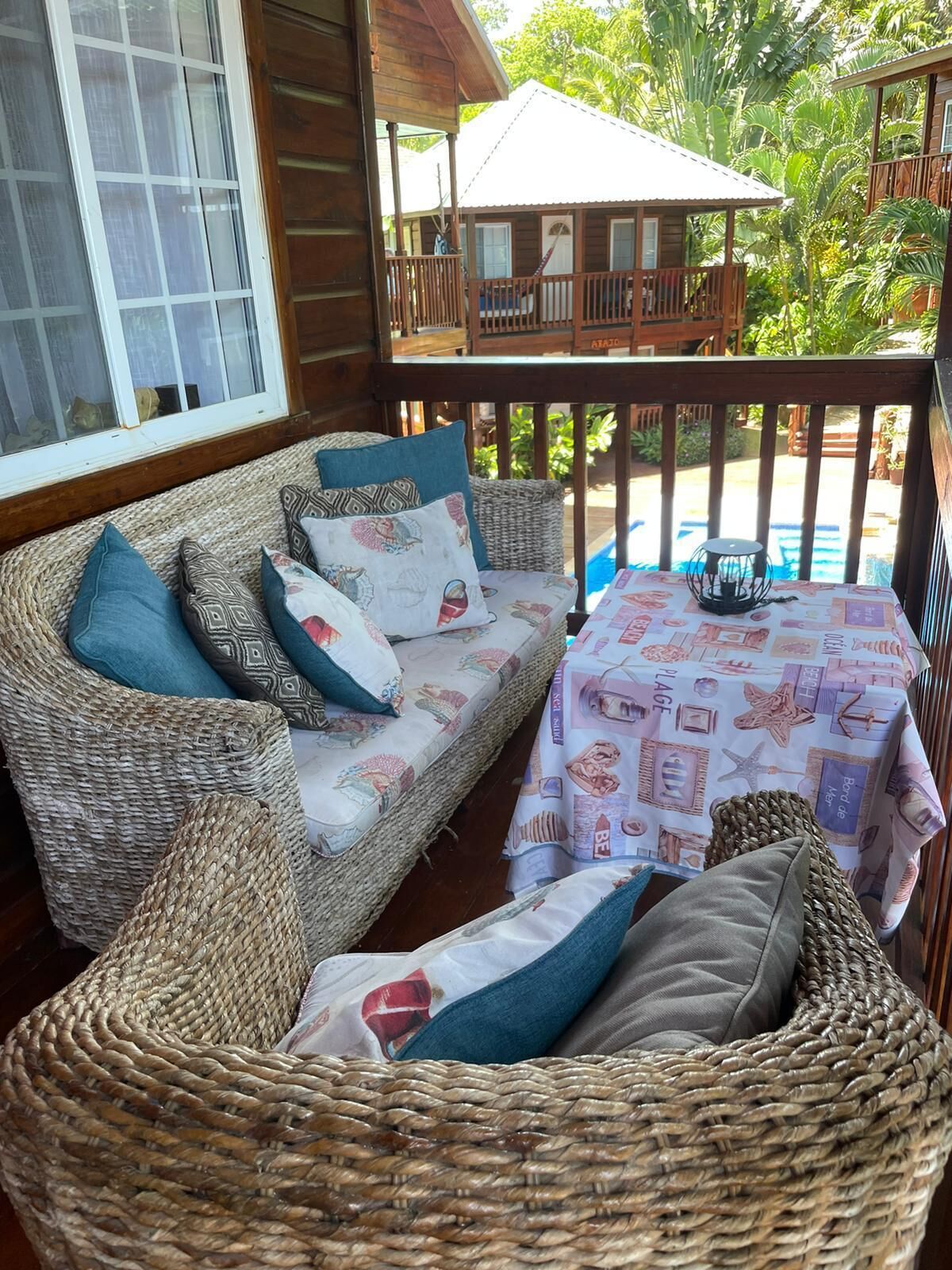 Un divano in vimini con cuscini decorati, un tavolino con tovaglia a tema spiaggia e vista su piscina e vegetazione lussureggiante.