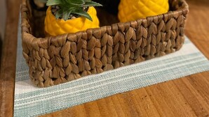 A woven basket containing two yellow bell peppers with green tops, placed on a wooden surface with a striped cloth underneath.