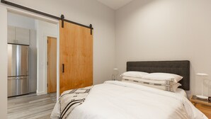 Un dormitorio con cama, una mesita de noche, una lámpara, una puerta corredera de madera y una zona de cocina con refrigerador.
