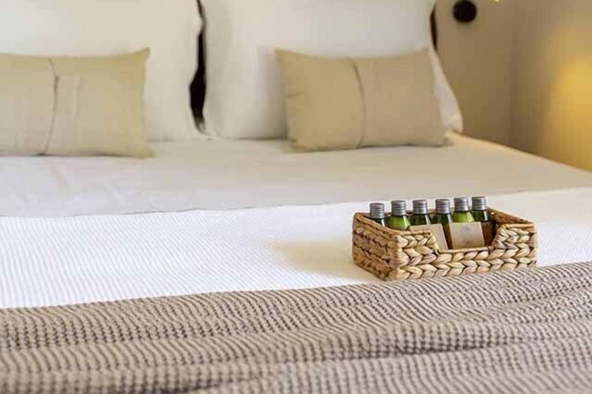 Una cama con sábanas blancas y almohadas color beige, una cesta tejida con botellas y un adorno en la pared.
