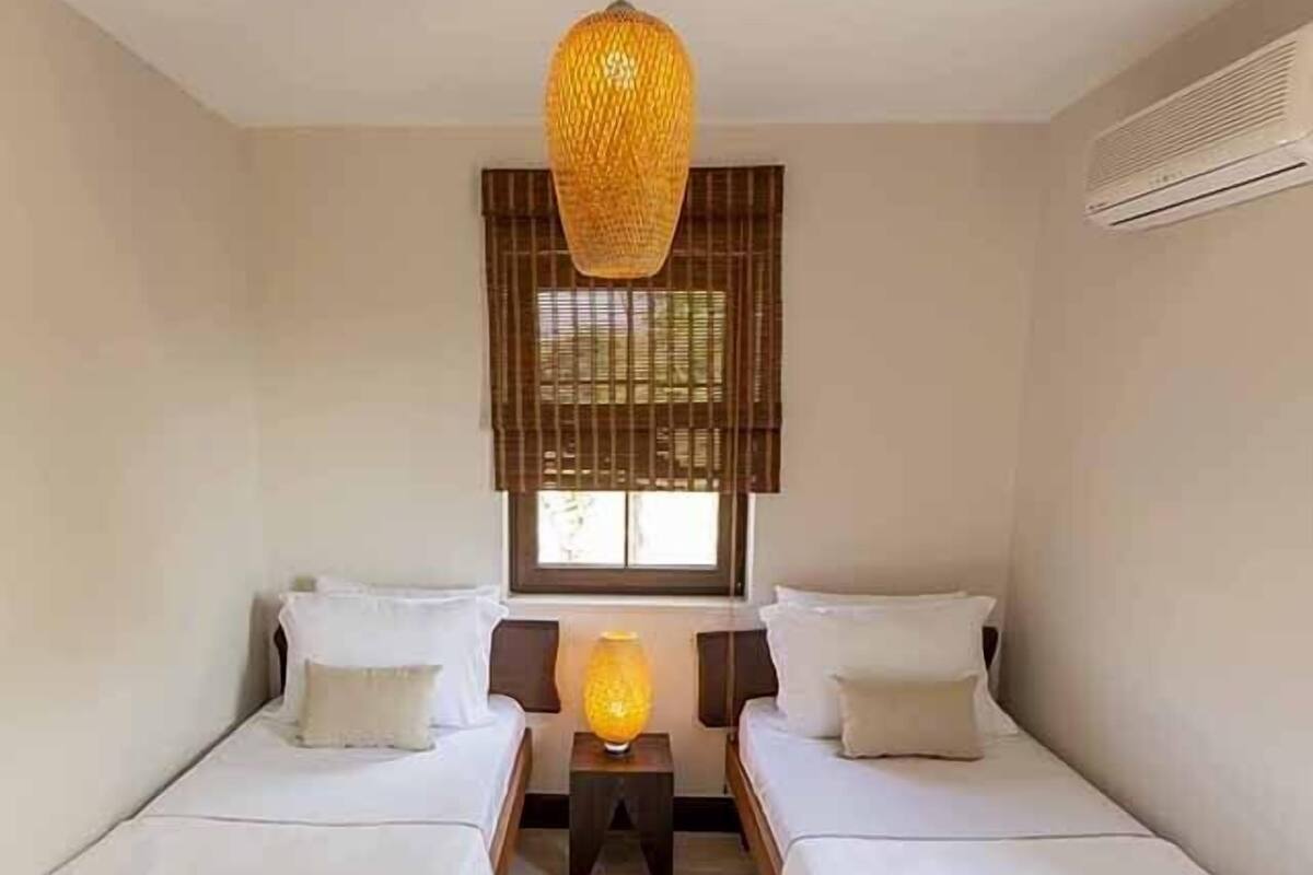 Una habitación con dos camas individuales, una lámpara colgante, una ventana con persianas y una pequeña mesa con lámpara.