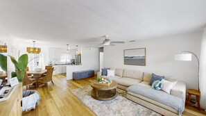 A spacious living area with a sofa, coffee table, and dining set. There is a kitchen in the background and a ceiling fan. - Beach Access + Coastal + 2 KING beds + Brand New: “Sunset on Madeira” (Madeira Beach)