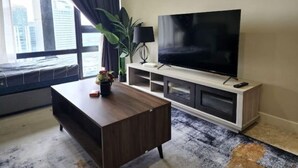 Una sala moderna con un televisor de pantalla plana grande sobre un soporte de madera, un sofá de color oscuro, una mesita de centro y una planta en maceta.
