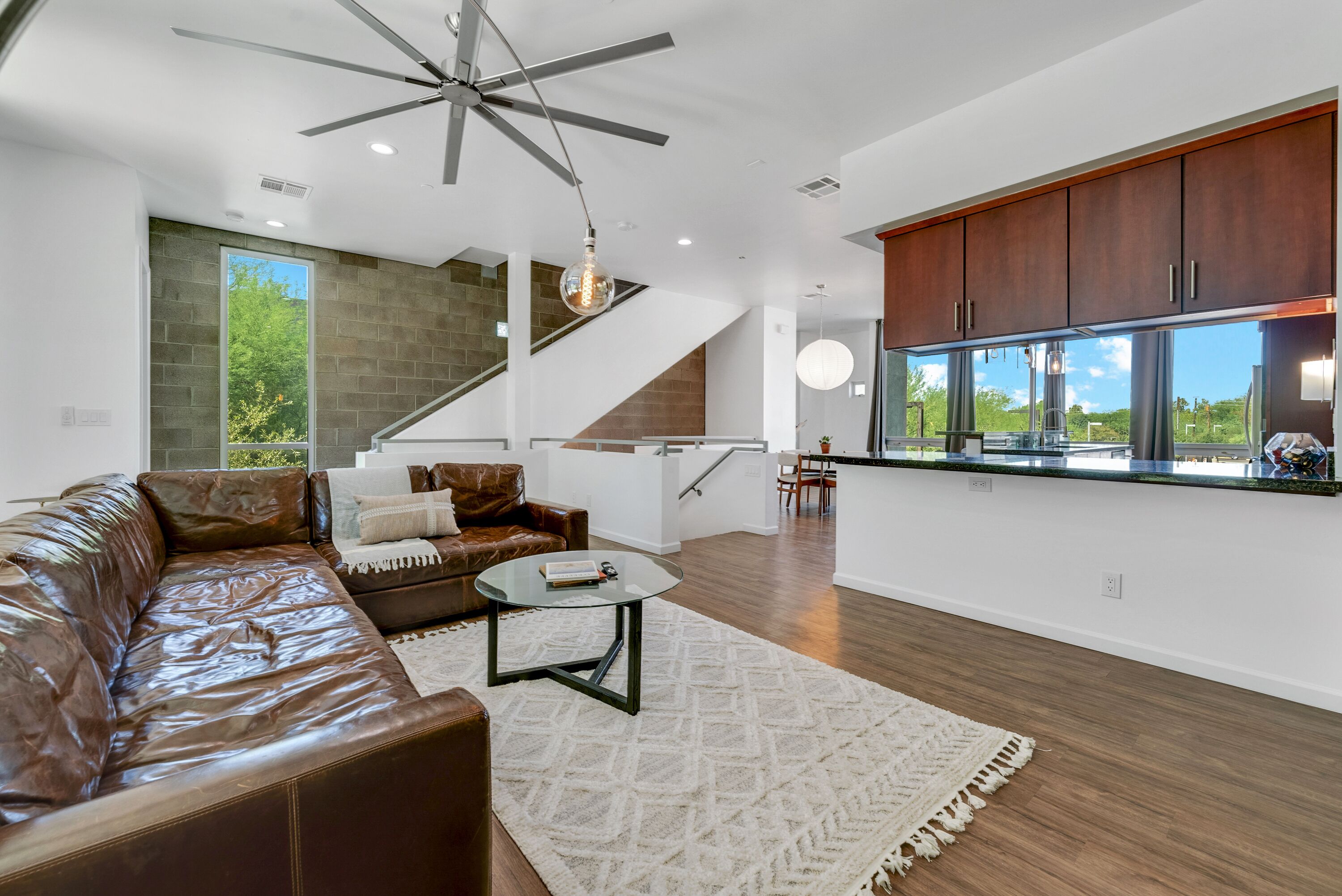 A modern living room with a leather couch, a glass coffee table, and a staircase leading to a kitchen with wooden cabinets and a large window.