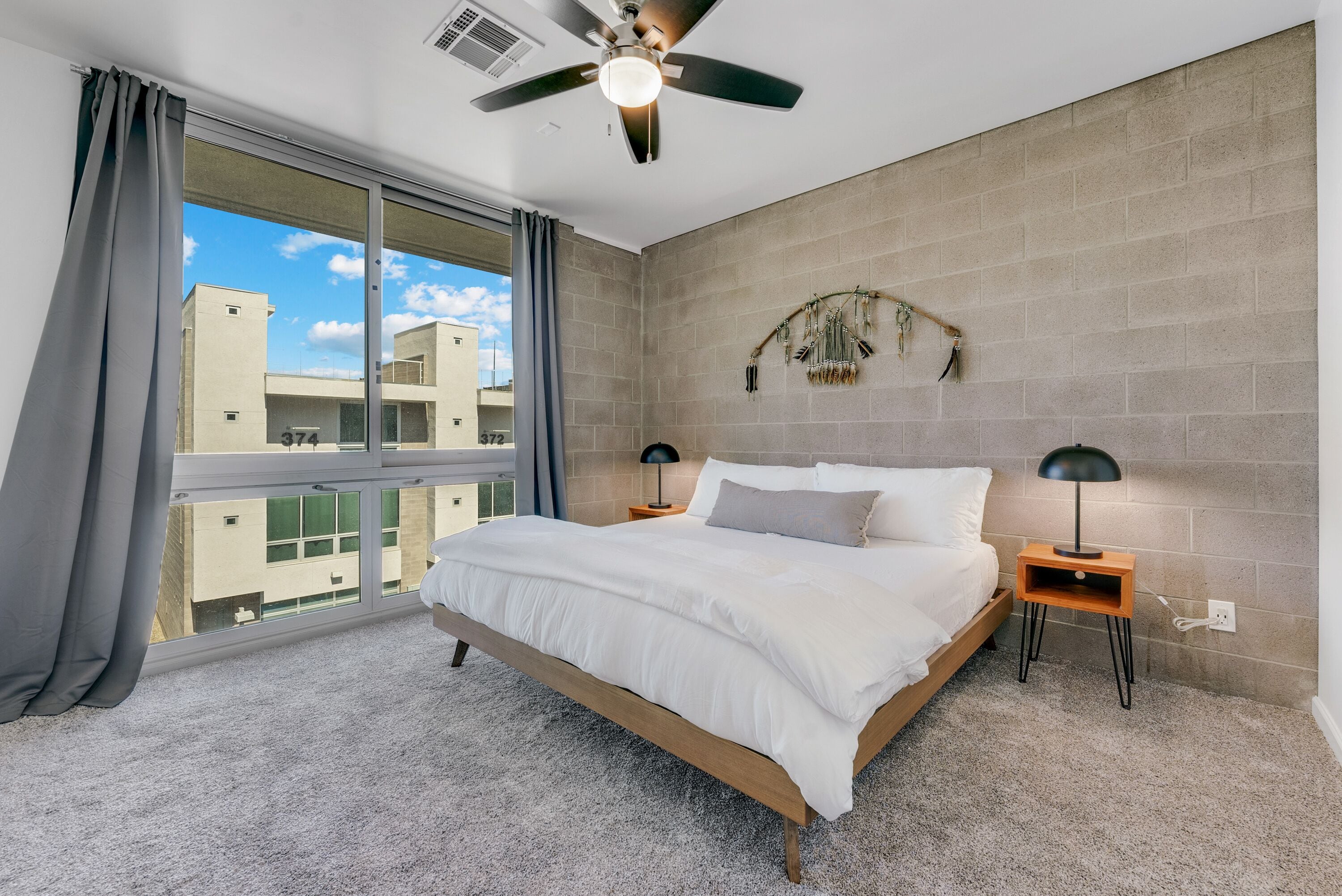 A bedroom with a large bed, two bedside tables, a ceiling fan, and a view of buildings through a window.