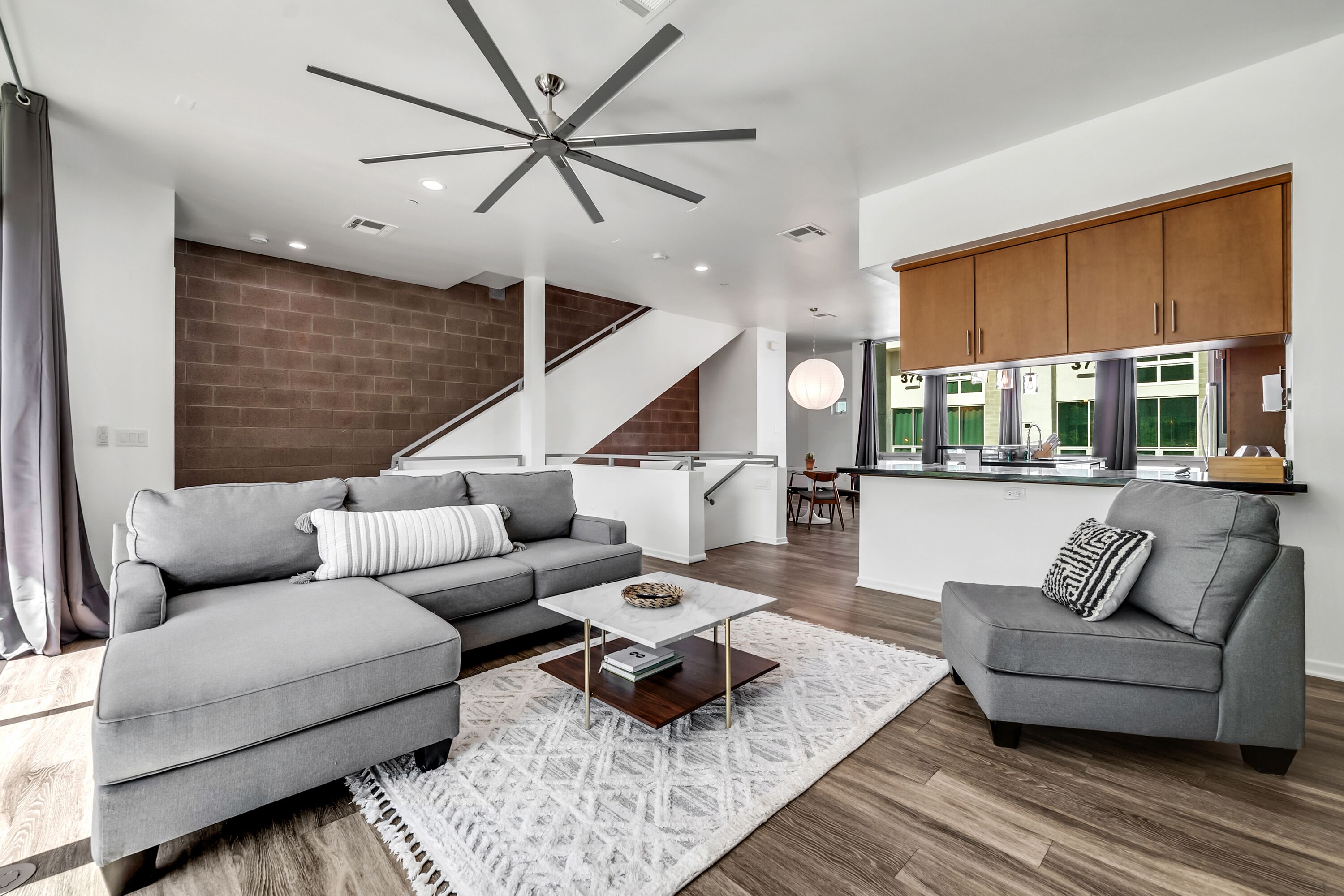 A modern living room with a grey sectional sofa, a coffee table, and a grey armchair. A staircase leads to a kitchen area with wooden cabinets and a dining table.