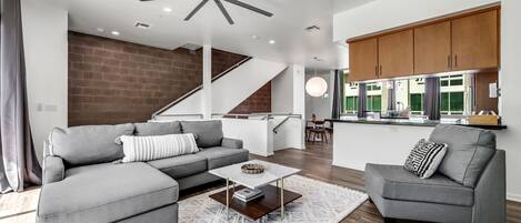 A modern living room with a grey sectional sofa, a coffee table, and a grey armchair. A staircase leads to a kitchen area with wooden cabinets and a dining table.