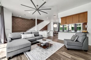 A modern living room with a grey sectional sofa, a coffee table, and a grey armchair. A staircase leads to a kitchen area with wooden cabinets and a dining table. - Tempe Lofts (Tempe)