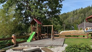 A wooden playground with a slide and swings, surrounded by a gravel area and greenery. - Family vacation home with 6 studio apartments. (Ceský Jiretín)