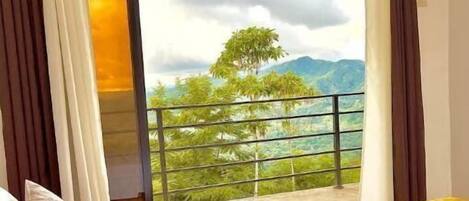 A bedroom with a bed, a view of a balcony with a railing, and a door leading to an outdoor area with trees and mountains.