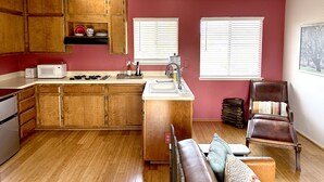 A compact kitchen with wooden cabinets, a microwave, and a stove. There are two windows with blinds, a sink, and a chair with a cushion.
