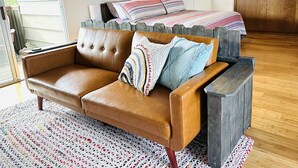 A cozy living room with a brown leather sofa, a colorful woven rug, and a bed with striped bedding.