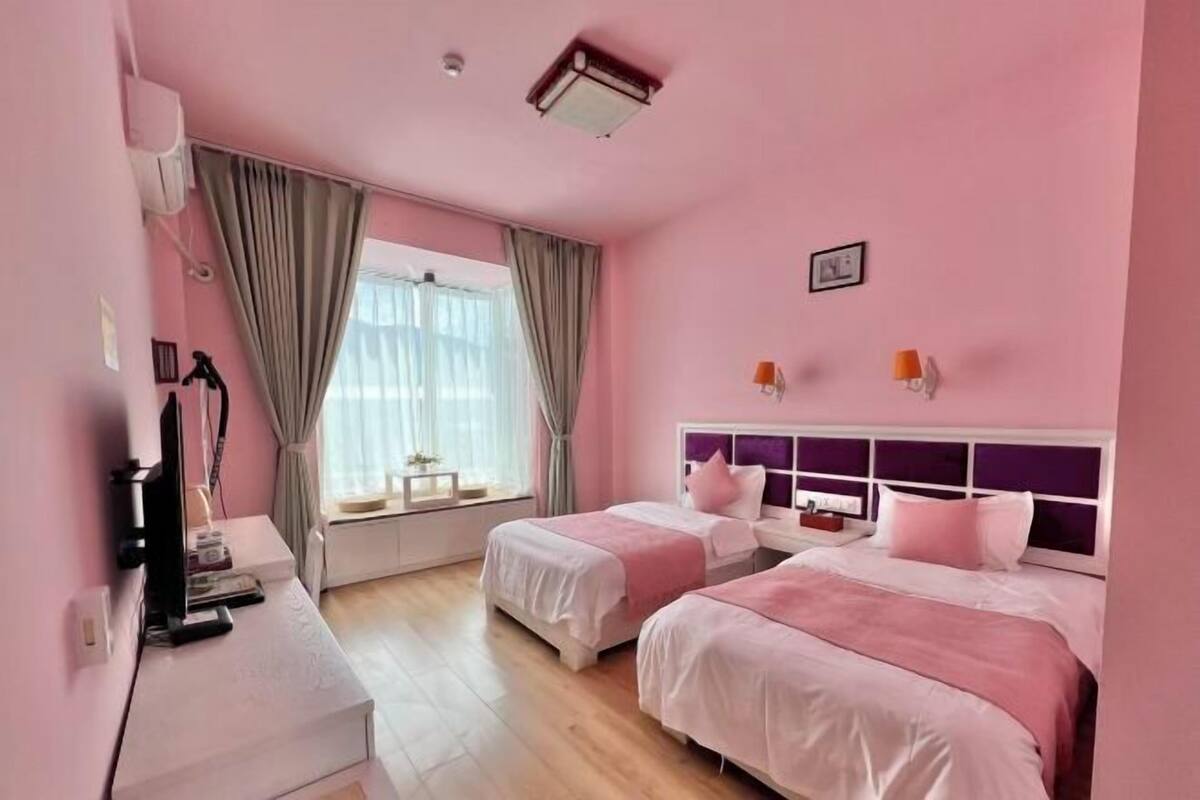 Une chambre d’hôtel décorée dans des tons roses, avec deux lits, un bureau et une fenêtre avec des rideaux.
