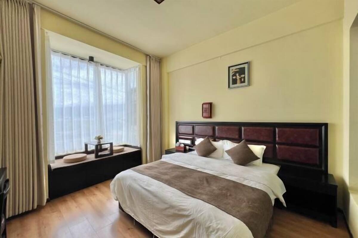Une chambre d’hôtel avec un grand lit, une fenêtre avec des rideaux, une table de chevet et un tableau encadré au mur.
