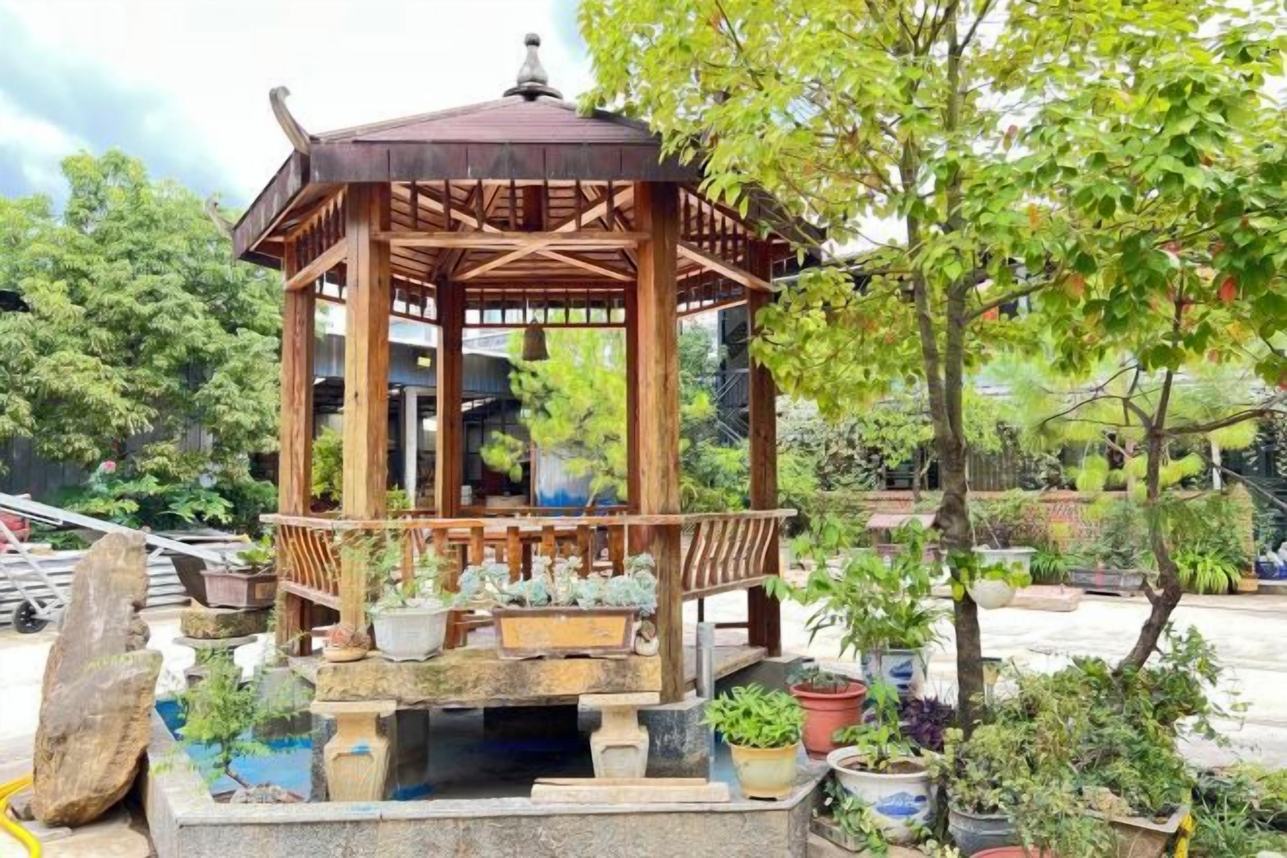 Un gazebo in legno con tetto, circondato da piante in vaso e una base in pietra.