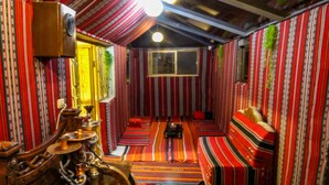 Ein schmaler Raum mit gestreifter Wandgestaltung und einem grünen Teppich. Es steht eine hölzerne Bank mit Kissen sowie ein kleiner Tisch mit Lampe und einem dekorativen Gegenstand darin.